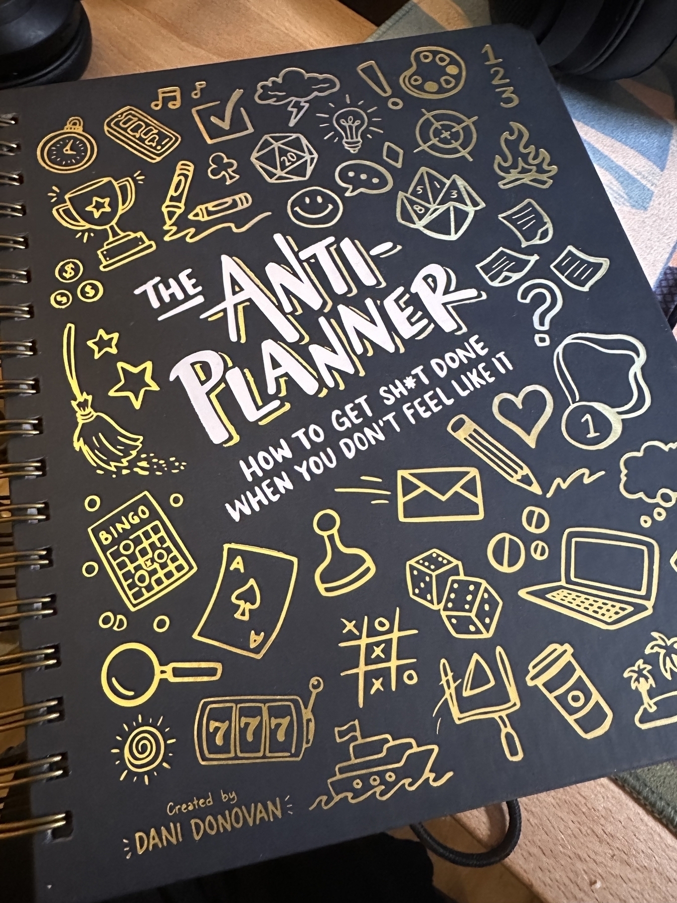 The cover of The Anti-Planner: How to Get Shit Done When You Don’t Feel Like It by Dani Donovan. It is a spiral bound book with navy background and hand drawn sketches of various doodles done in gold foil.